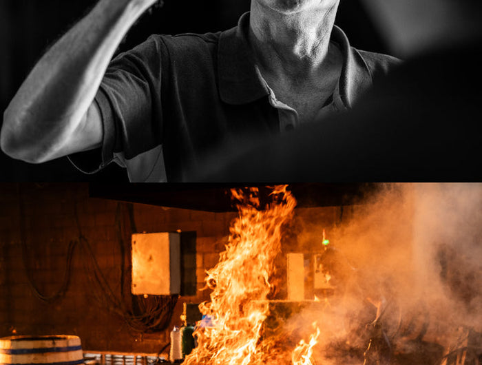 Man holding a glass of whisky above an image of whisky barrels on fire