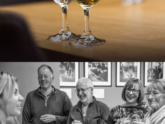 Two wine glasses filled with a whisky on a wooden table, with people socializing in the background.